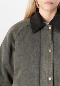 Giacca di jeans verde con colletto in corduroy nero, chiusura con bottoni a pressione, cuciture visibili e vestibilità rilassata, che mette in evidenza un tessuto testurizzato.