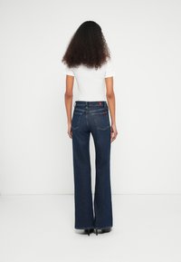 Jean évasé bleu foncé à taille haute, avec poches arrière et une étiquette rouge. Associé à un t-shirt blanc uni, vu de dos.