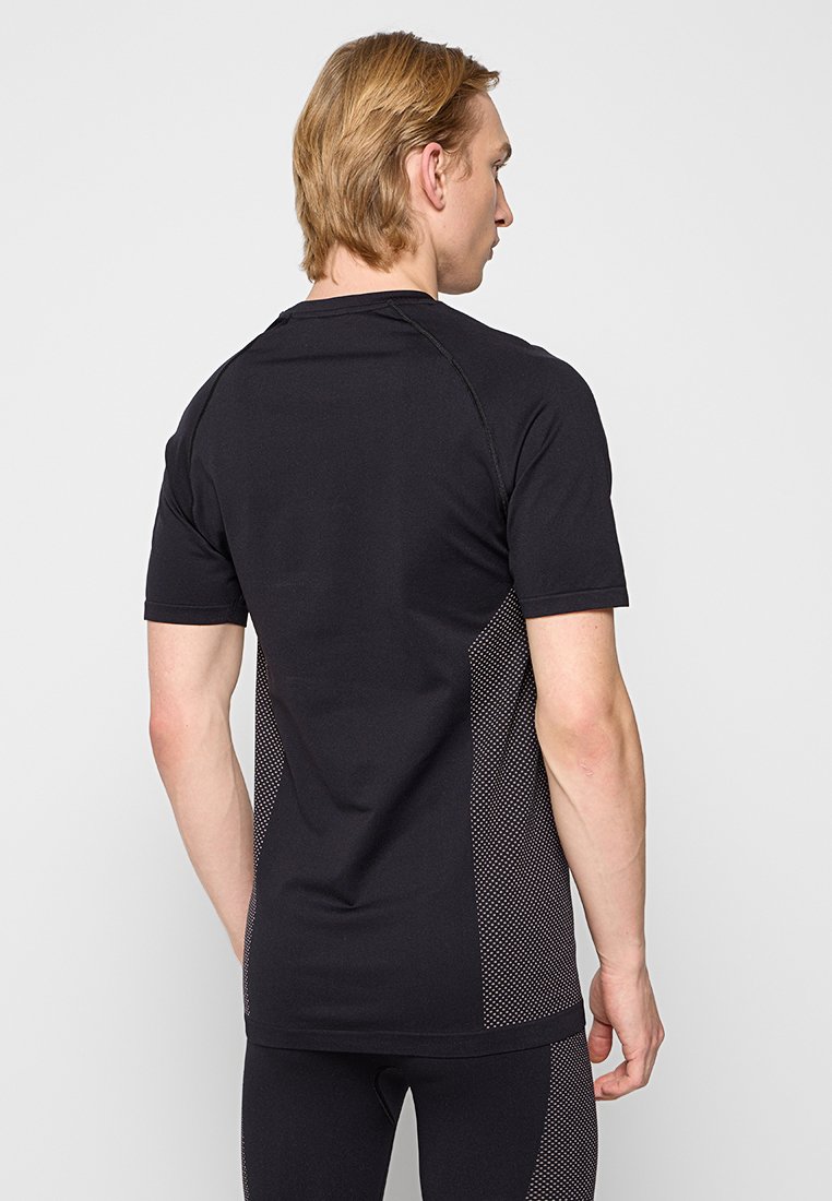 Mann trägt ein schwarzes, kurzärmliges Sportshirt und passende Leggings mit gepunkteten Seitenpanels, steht mit dem Rücken zu einem schlichten, hellen Hintergrund.