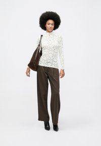 Mujer con peinado afro, vistiendo una blusa de encaje blanco, pantalones de pierna ancha marrones, botas negras y llevando un gran bolso de hombro marrón, de pie sobre un fondo blanco.