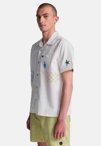 Joven con una camisa blanca de manga corta y botones con parches gráficos y pantalones cortos verde claro sobre un fondo simple.