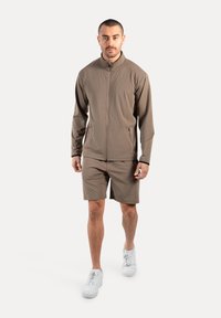 Sportliches Herrenbekleidungsset bestehend aus einer taillierten, braunen Jacke mit Reißverschluss und passenden Shorts, gefertigt aus leichtem, glattem Material. Weiße Sneakers runden den Look ab.