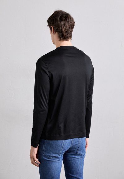 T-shirt noir à manches longues en tissu lisse avec un col rond. Porté avec un jean en denim bleu, affichant un design simple au dos.