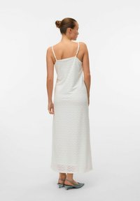 Maxi jurk in het wit, met een zigzagpatroon, dunne bandjes en een vloeiende silhouet. Gecombineerd met lichtblauwe hoge hakken.