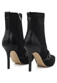 Stivaletti neri alla caviglia in una combinazione di suede e pelle lucida, punta affilata, alto tacco a stiletto e cinturino alla caviglia con dettagli a catena.