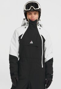 Mono de esquí con un diseño en blanco y negro, que cuenta con un cuello alto, un bolsillo frontal, cierres de cremallera y puños ajustables. El material parece ser impermeable.