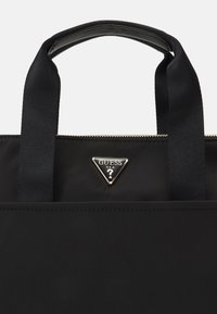Czarna torba na ramię marki Guess z dwoma uchwytami, złotym zamkiem błyskawicznym oraz trójkątną metalową naszywką z logo umieszczoną na środku poniżej zamka.