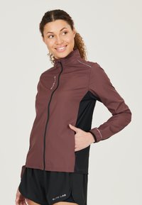 Veste de sport bordeaux et noire avec un col montant, fermeture éclair et détails réfléchissants, fabriquée en tissu léger et respirant.