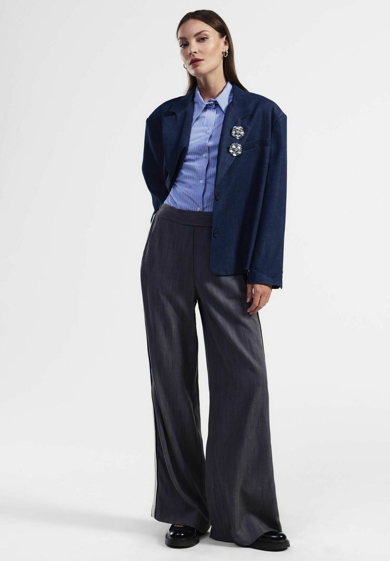 Giacca blu navale con ampi reverse, spille a forma di fiore in argento, abbinata a pantaloni grigi a gamba larga con finitura testurizzata e scarpe nere.