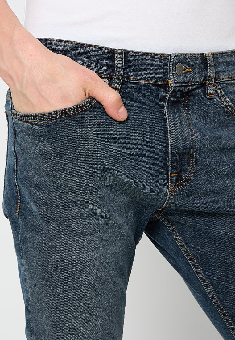 Jean en denim bleu foncé, à la coupe slim, avec une conception à cinq poches et des accents de couture orange. Texture lisse avec une fermeture à bouton classique.