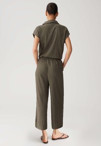 Olive green jumpsuit with short sleeves, textured fabric, elastic waistband, and wide-leg design. Features a back pocket and flat sandals.
