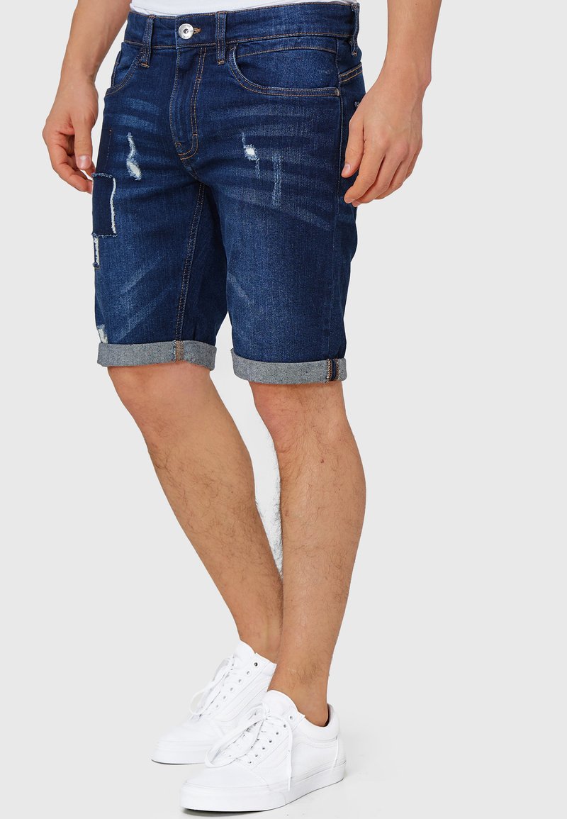 Dunkelblaue Jeansshorts mit Used-Effekten, umgeschlagenen Bündchen und Fünf-Taschen-Design, kombiniert mit weißen Sneakern.