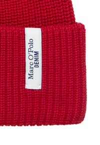 Czerwona dzianinowa czapka z prążkowaną fakturą, z białą etykietą z niebieskim napisem "Marc O'Polo DENIM" przyszytą z boku.