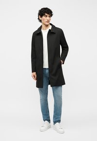 Νέος άνδρας που φοράει μαύρο παλτό πάνω από λευκό πουκάμισο, μπλε τζιν και λευκά αθλητικά παπούτσια, στέκεται με το ένα χέρι στην τσέπη του παλτού.