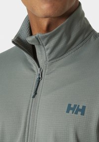 Chaqueta de cremallera verde claro con un alto cuello, forro interior texturizado, costuras planas y un pequeño logotipo en el pecho. Acabado de tela suave.