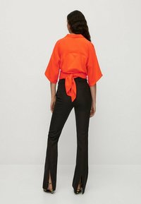 Blouse orange fluide avec un détail noué à la taille, associée à un pantalon noir ajusté avec des fentes latérales à l'ourlet. Texture de tissu lisse.