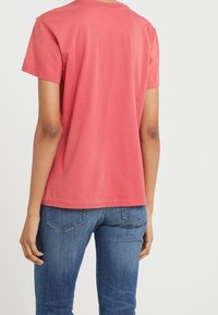Červené bavlněné tričko s krátkým rukávem, kulatým výstřihem a volným střihem; nošeno s modrými džínami s kapsami na zadní straně.