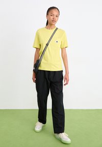 Žlté bavlnené tričko s vreckom na hrudi, skombinované s čiernymi voľnými nohavicami a svetlými teniskami, vytvárajúce neformálny outfit.