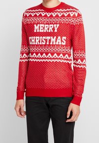 Rode gebreide trui met witte "MERRY CHRISTMAS" tekst, geometrische patronen, en geribbelde boorden en zoom. Gestructureerde stof met een feestelijk thema.