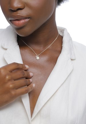 Zilveren ketting met een gestructureerd, rond hangertje en een klein accentkraaltje, gedragen boven een lichtgekleurde blouse, tegen een neutrale achtergrond.
