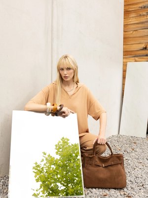 Blonde woman in beige outfit kneels on gravel, holding a large brown handbag and a mirror reflecting green tree leaves.