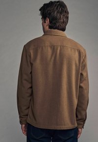 Brun, tekstureret jakke med lige snit, knappede manchetter og klassisk krave. Båret med blå jeans, set fra ryggen.