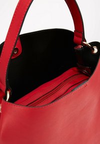 Červená syntetická kabelka s černým interiérem, vybavená zipovou kapsou a dvojitými úchyty. Hladký povrch a zlaté kovové akcenty.