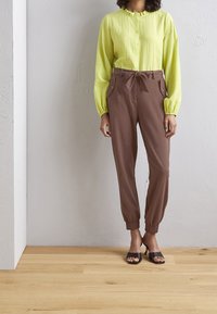 Kvinna iklädd en limegrön långärmad blus instoppad i högmidjade bruna tapered-byxor med ett knytbälte och svarta öppna klackskor.