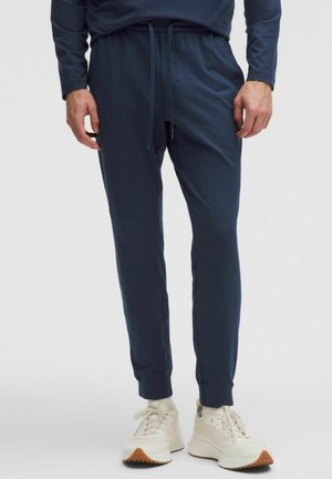 Granatowe spodnie dresowe z troczkiem w pasie, zwężanymi nogawkami i elastycznymi ściągaczami, wykonane z gładkiego, elastycznego materiału. Noszone z jasnymi sneakersami.