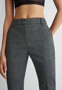 Kvinde iført grå rutede skrædderbukser med høj talje, en forkantfold og sidelommer, parret med en sort cropped top med blonder.