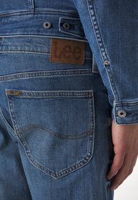 Vue arrière d'une personne portant un jean en denim bleu Lee et une veste en denim assortie avec des patchs et boutons visibles de la marque Lee.