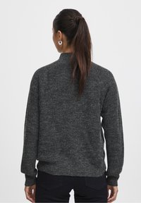 Frau mit langen, dunklen Haaren in einem Pferdeschwanz, die silberne Creolen trägt, einen dunkelgrauen Strickpullover und schwarze Hosen, von hinten gezeigt.
