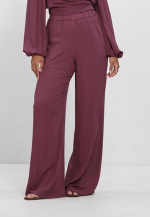 Vrouw draagt bordeauxrode broek met hoge taille en wijd uitlopende pijpen, gecombineerd met bijpassende top met lange mouwen, staand op een witte vloer in sandalen.