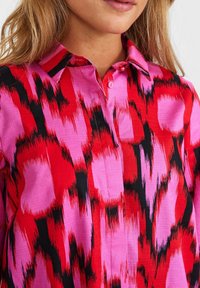 Camicetta a maniche corte in un vivace motivo rosa, rosso e nero. Presenta un colletto classico e chiusura con bottoni, con una texture liscia e un design audace.