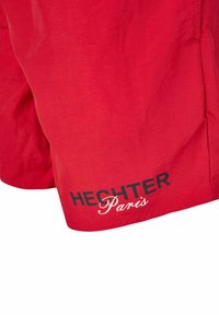 Shorts de bain rouges en tissu léger. Présente un texte noir et blanc indiquant "HECHTER" et "Paris" sur l'ourlet inférieur.