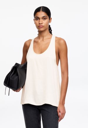 Kobieta z warkoczami, ubrana w kremowy top bez rękawów i czarne spodnie, trzymająca w lewej ręce czarną torbę ze ściągaczem.