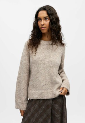 Light beige knit sweater with a relaxed fit, round neckline, and ribbed cuffs. Worn over a brown plaid skirt, featuring soft, textured fabric.