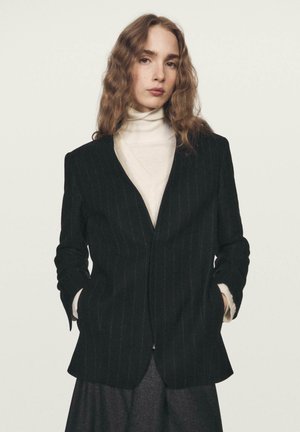 Blazer a righe sottili nere in misto lana, con scollo a V profondo, due tasche frontali e maniche lunghe. Abbinato a un dolcevita bianco.