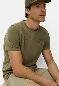 Mand iført olivengrøn kasket, olivengrøn t-shirt med brystlomme og beige shorts, siddende og kiggende til siden mod en hvid baggrund.