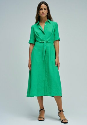 Mujer con cabello largo y oscuro que lleva un vestido camisa midi verde con la cintura atada y sandalias negras de tacón alto con tiras, de pie contra un fondo liso.