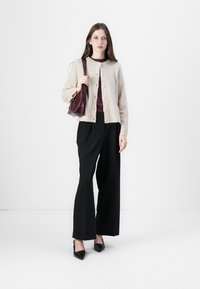 Licht beige gebreide cardigan met knopen, bordeaux top, zwarte wijde broek en zwarte puntige hakken. Houdt een kleine bordeaux handtas vast.