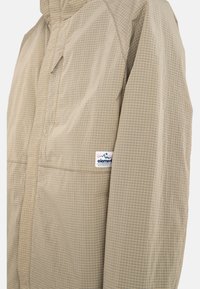 Chaqueta ligera beige con un patrón de cuadrícula, con cierre frontal de cremallera y un parche de logo en la manga izquierda. Textura suave, corte relajado.