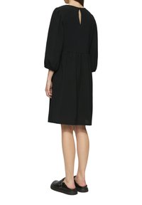 Schwarzes Kleid mit dreiviertellangen Puffärmeln, Detail mit Schlüsselloch am Rücken, geraffter Taille und einem leichten strukturierten Stoff. Kombiniert mit schwarzen Slippern.
