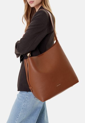 Frau trägt schwarzen Blazer und hellblaue Jeans, trägt eine große braune Ledertasche mit Textur auf der Schulter.