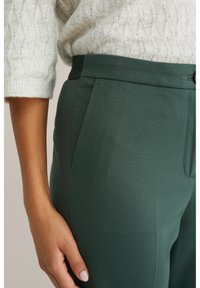 Személy zöld, magas derekú nadrágban és krémszínű, háromnegyedes ujjú kötött pulóverben, keze a combján pihen.