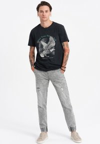 T-shirt graphique noir avec un motif d'oiseau, associé à un pantalon déchiré gris clair et des baskets beiges. Coupe décontractée avec une texture apparente.