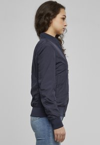 Jachetă bomber bleumarin cu guler mulat, fermoar frontal, buzunare laterale și manșete striate. Textură netedă, țesătură ușoar ă.