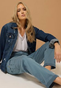 Jeansjacka med slitna detaljer, vit V-ringad t-shirt och ljusblå cargobyxor med fickor och uppvikta muddars, på en neutral bakgrund.