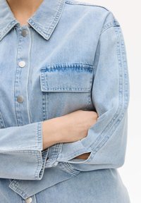 Giacca di jeans azzurro chiaro con collo, bottoni a pressione argentati, taschino sul petto, vestibilità leggermente ampia e polsini aperti.