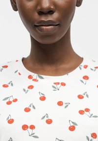 Κοντινό πλάνο ενός ατόμου που φοράει λευκό πουκάμισο με κόκκινο μοτίβο κερασιού και πράσινο φύλλο, παρουσιάζοντας το κάτω μέρος του προσώπου και τον λαιμό.